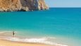 A beach with clear turquoise water, a person fishing, and rocky cliffs in the background.