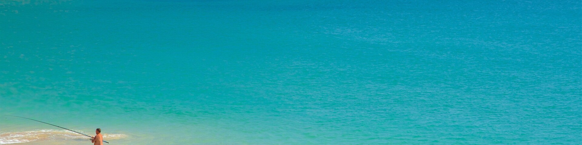 Una playa con agua turquesa cristalina, una persona pescando y acantilados rocosos al fondo.