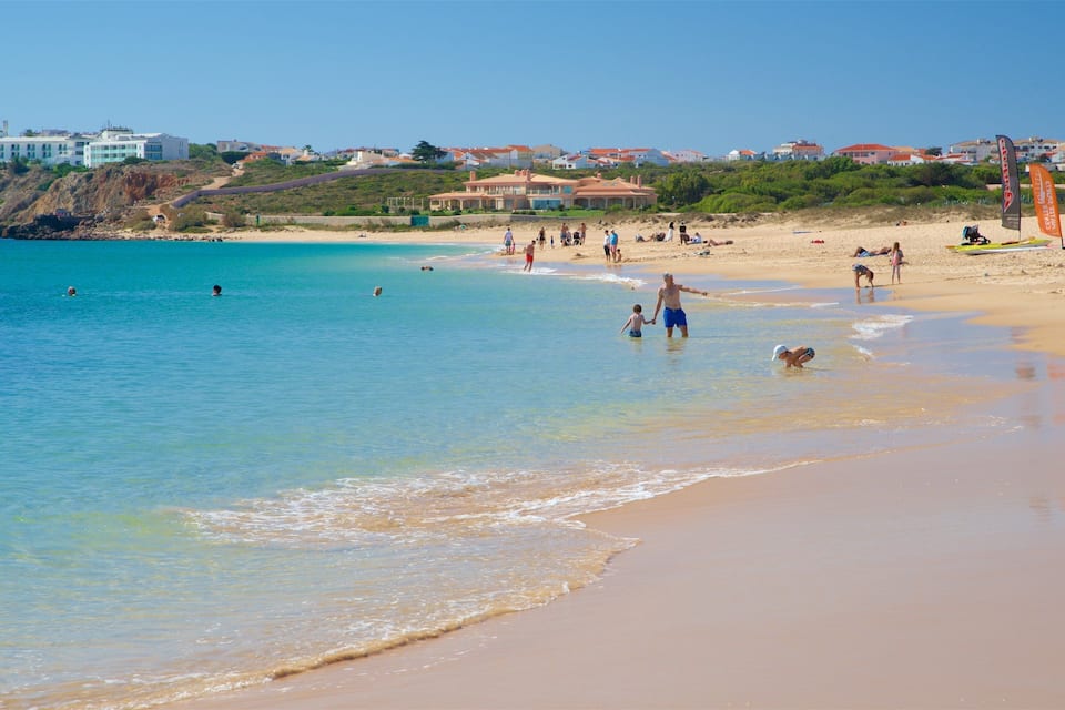 A beach with clear turquoise water, sandy shore, and houses in the background.
