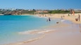 Een strand met helder turquoise water, een zandstrand en huizen op de achtergrond.