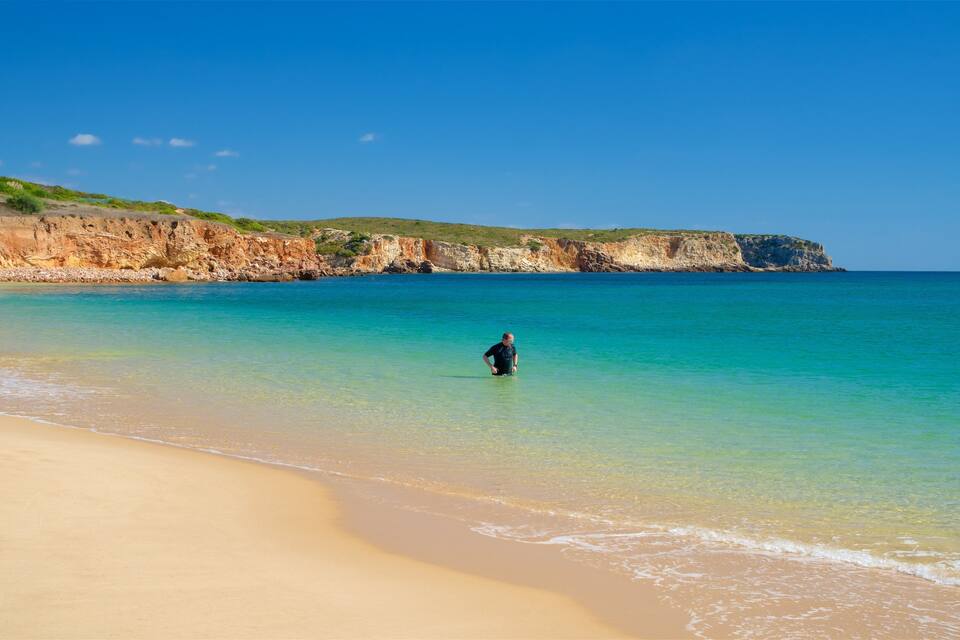 A beach with clear turquoise water, a sandy shore, and rocky cliffs in the background.