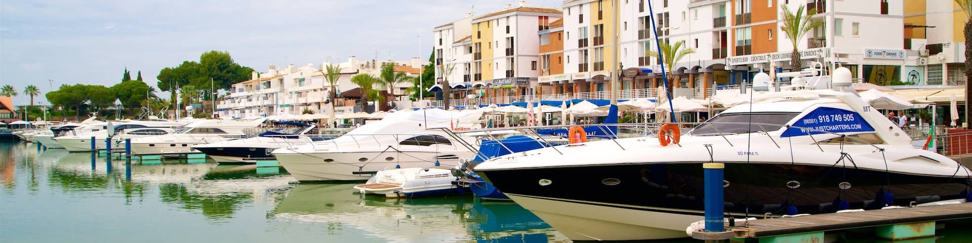 A marina with several yachts docked, surrounded by multi-story buildings with shops and restaurants.