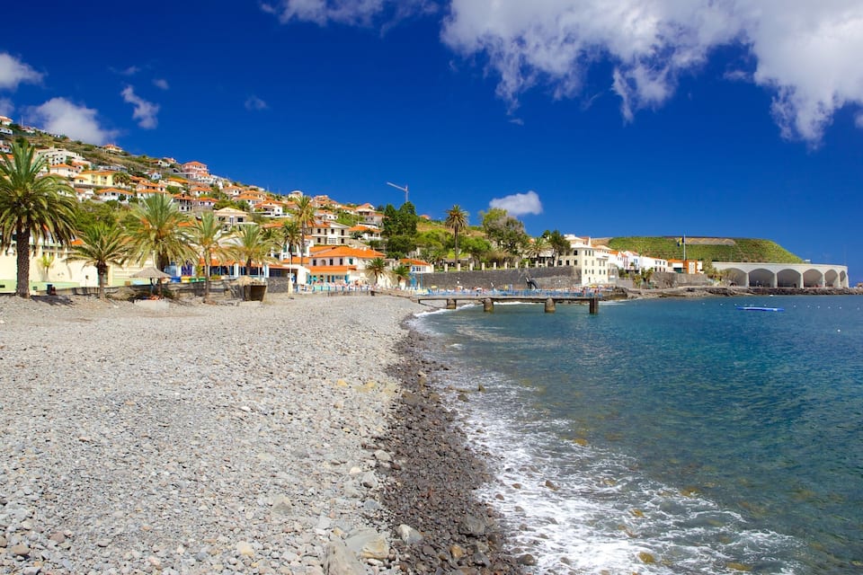 A pebble beach with a pier, palm trees, and colorful buildings on a hillside.