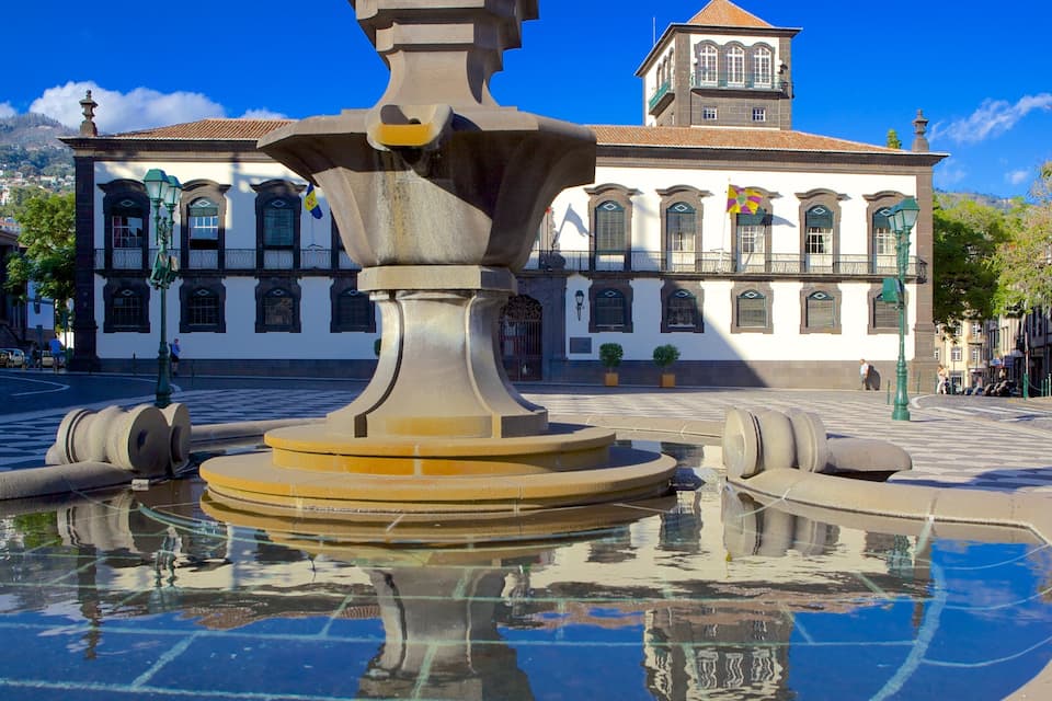 A historic fountain with a large basin in the center, surrounded by a cobblestone plaza. In the background, there is a white building with a tower and arched windows.