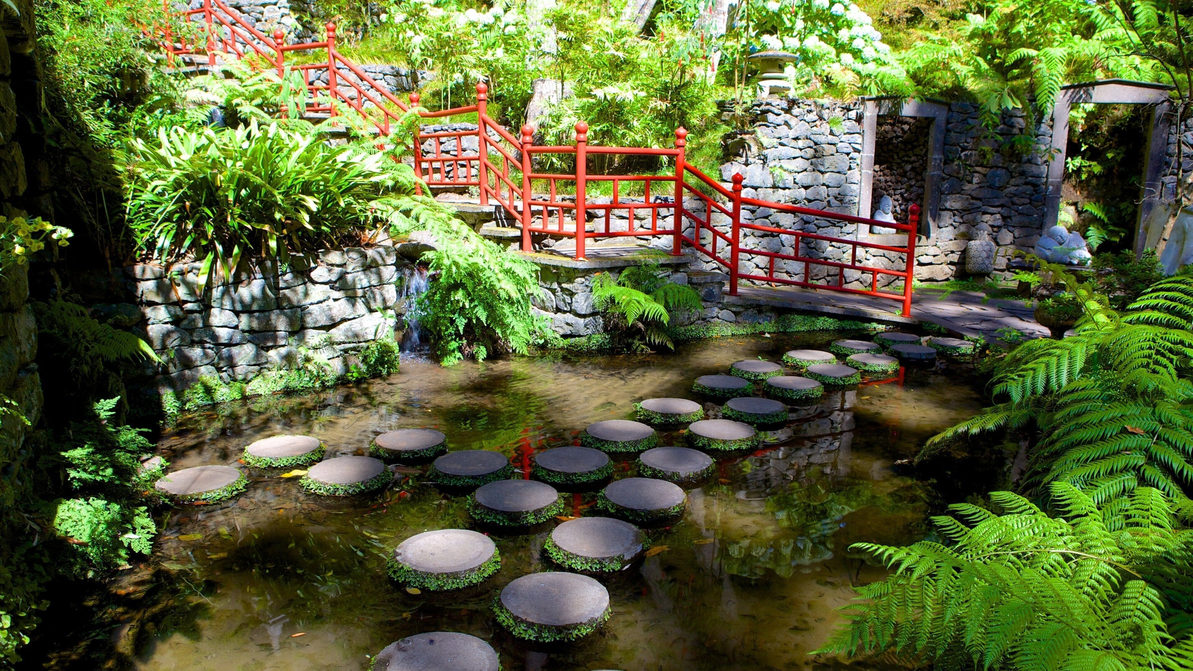 A garden with a stone bridge, red railing, and stepping stones over a stream.