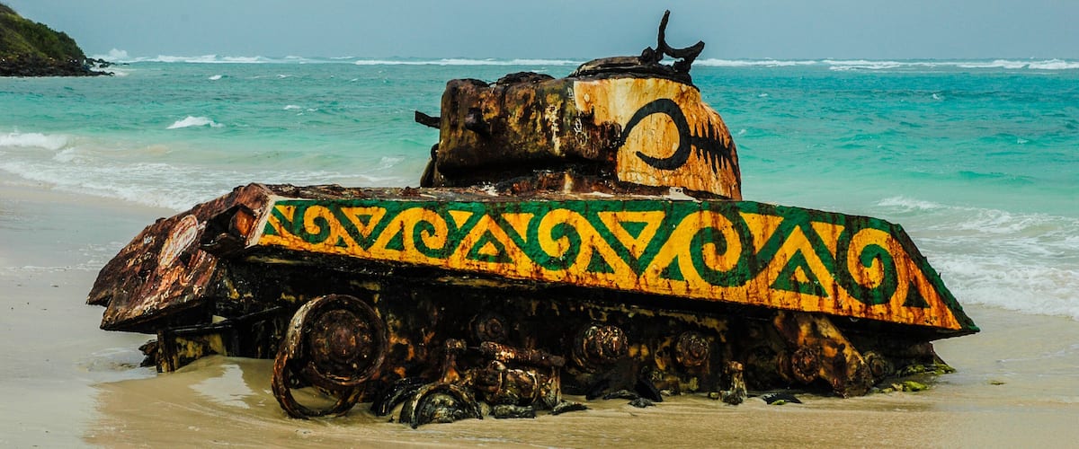 Um tanque militar enferrujado numa praia com água turquesa e céu nublado.