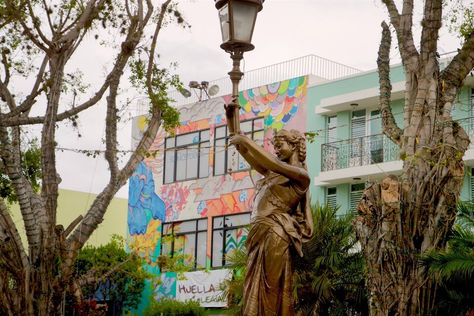 Estàtua d'una dona que sosté un fanal, envoltada d'arbres i un mural colorit a un edifici.