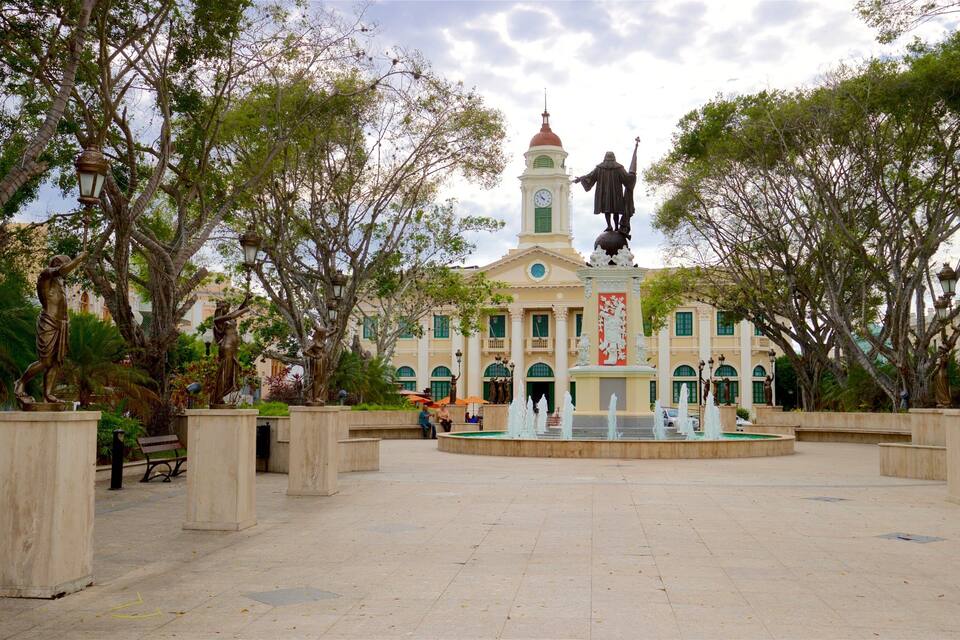A plaza with a fountain, a clock tower, and statues.