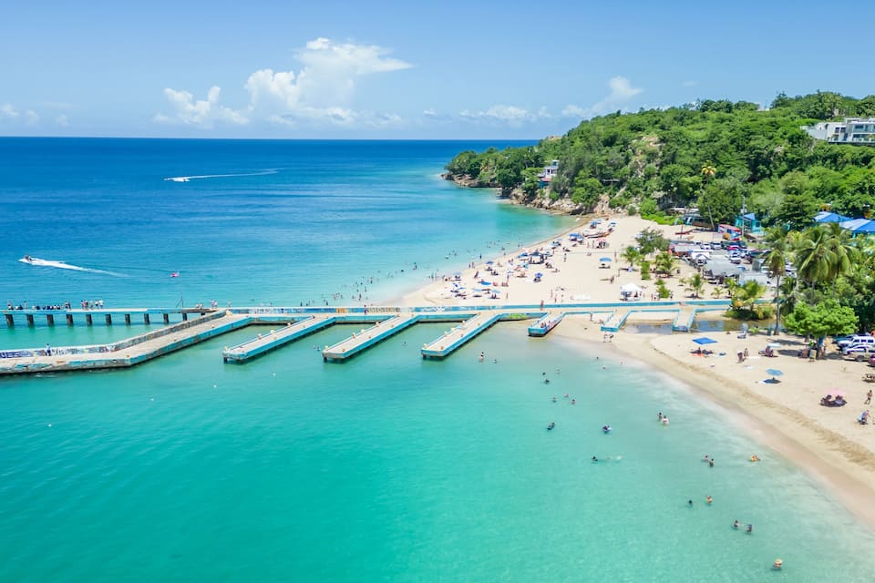 A beach with clear turquoise waters, a pier extending into the sea, and a hillside with buildings and palm trees.