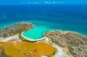Luftaufnahme einer Küstenlandschaft mit klarem türkisblauem Wasser, einem Sandstrand und einem deutlichen, gelblich gefärbten Gewässer.
