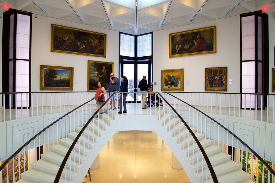 A grand staircase with white railings and a polished floor, flanked by large classical paintings.