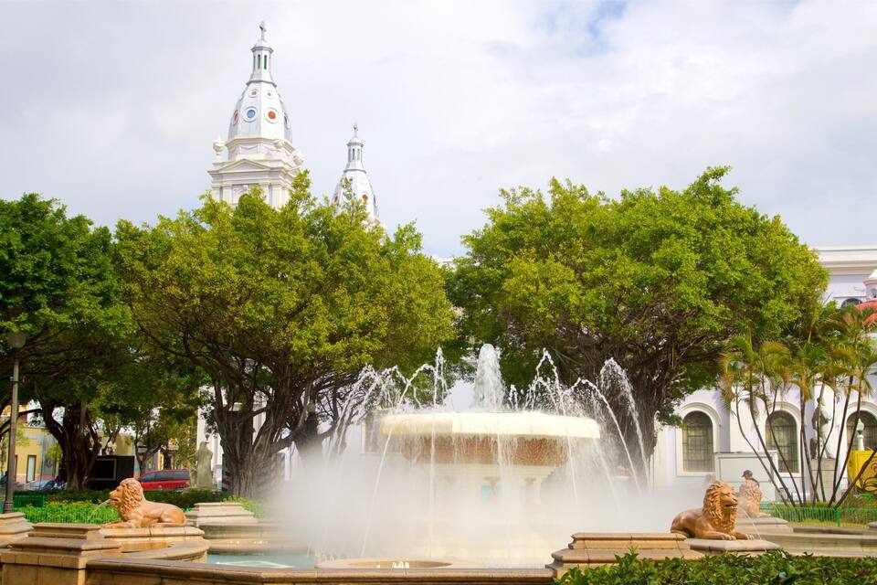 A fountain with lion statues, surrounded by trees and a white building with domes in the background.