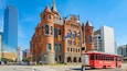 A historic building with red brick architecture and multiple towers, located at a city intersection with modern skyscrapers in the background.