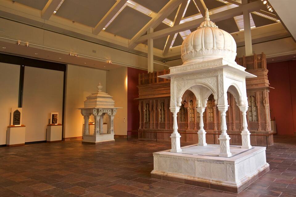 Um pavilhão branco ornamentado com esculturas detalhadas e teto abobadado, rodeado por um salão amplo com pé-direito alto e ladrilhos estampados.
