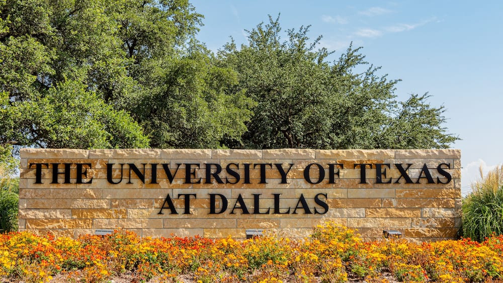 A sign for The University of Texas at Dallas with a backdrop of trees and colorful flowers.