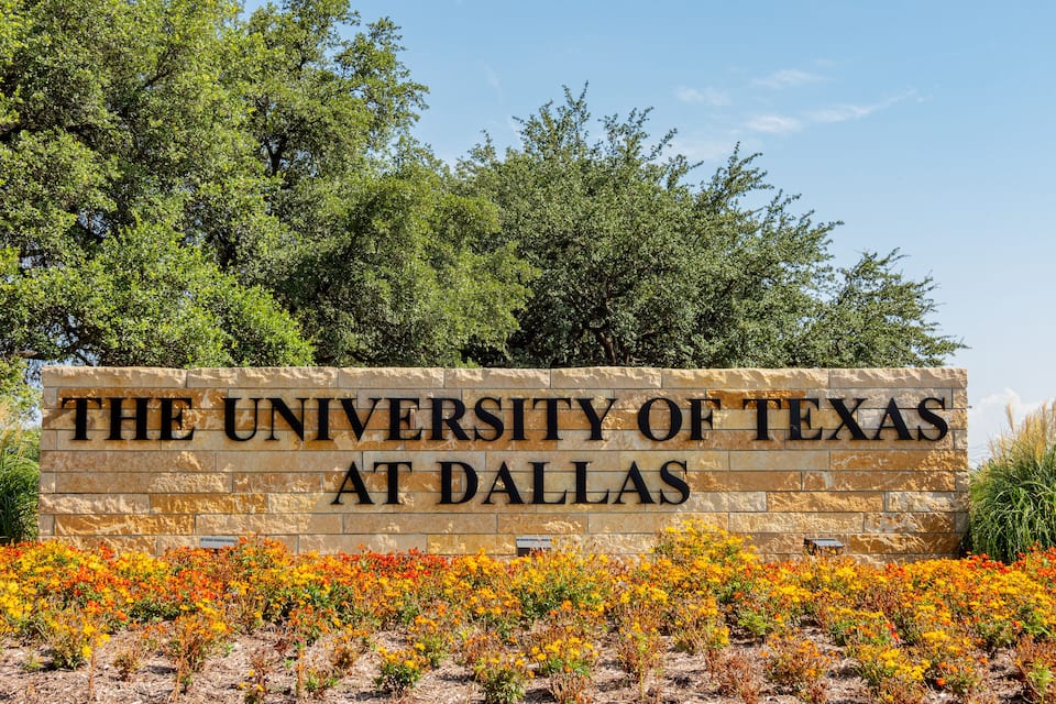 A sign for The University of Texas at Dallas with a backdrop of trees and colorful flowers.