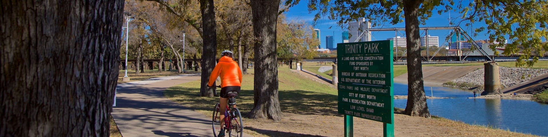 Ciclista en un camino pavimentado en el Parque Trinity, con un letrero que indica que es un área de conservación de tierra y agua.
