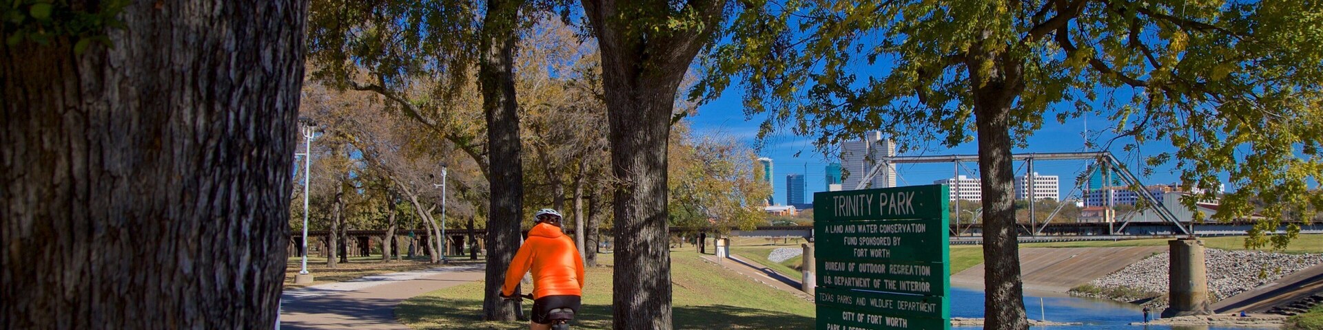 Ciclista en un camino pavimentado en el Parque Trinity, con un letrero que indica que es un área de conservación de tierra y agua.