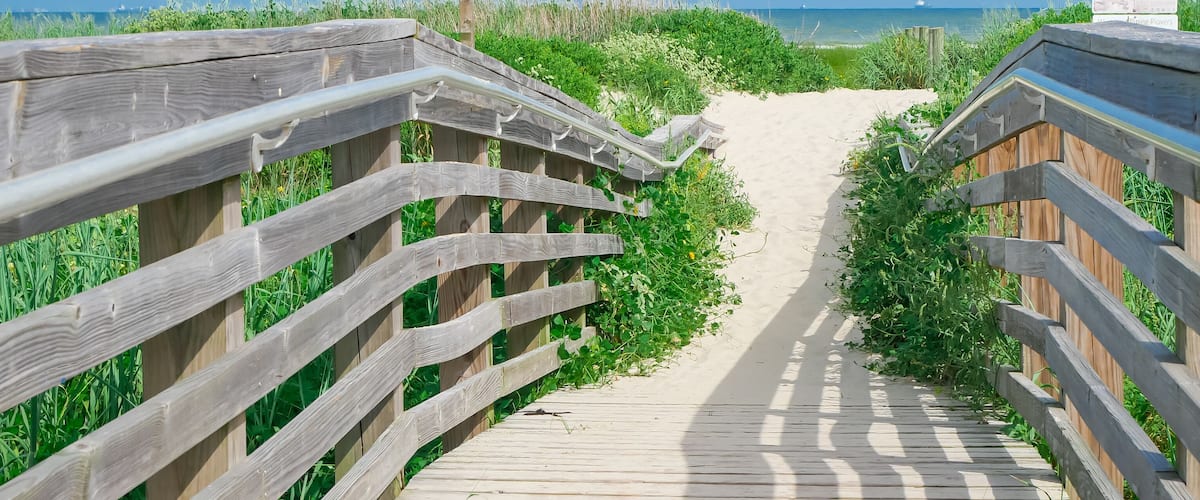 Panorama view boardwalk leads through coastal grass toward East Beach in Galveston, Texas. Elevated with railings on both sides, guiding the viewer eye toward the horizon under clear blue sky
