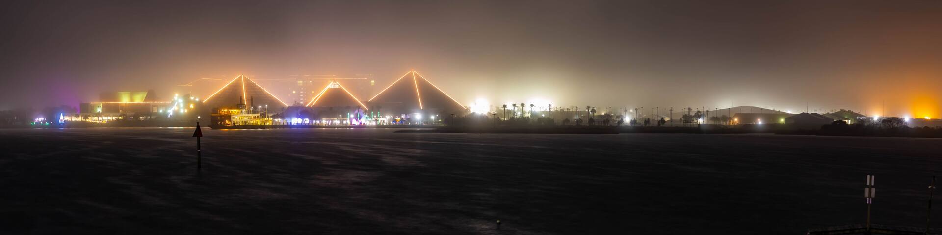 Vista nocturna de un frente marítimo con estructuras iluminadas y un ambiente brumoso.