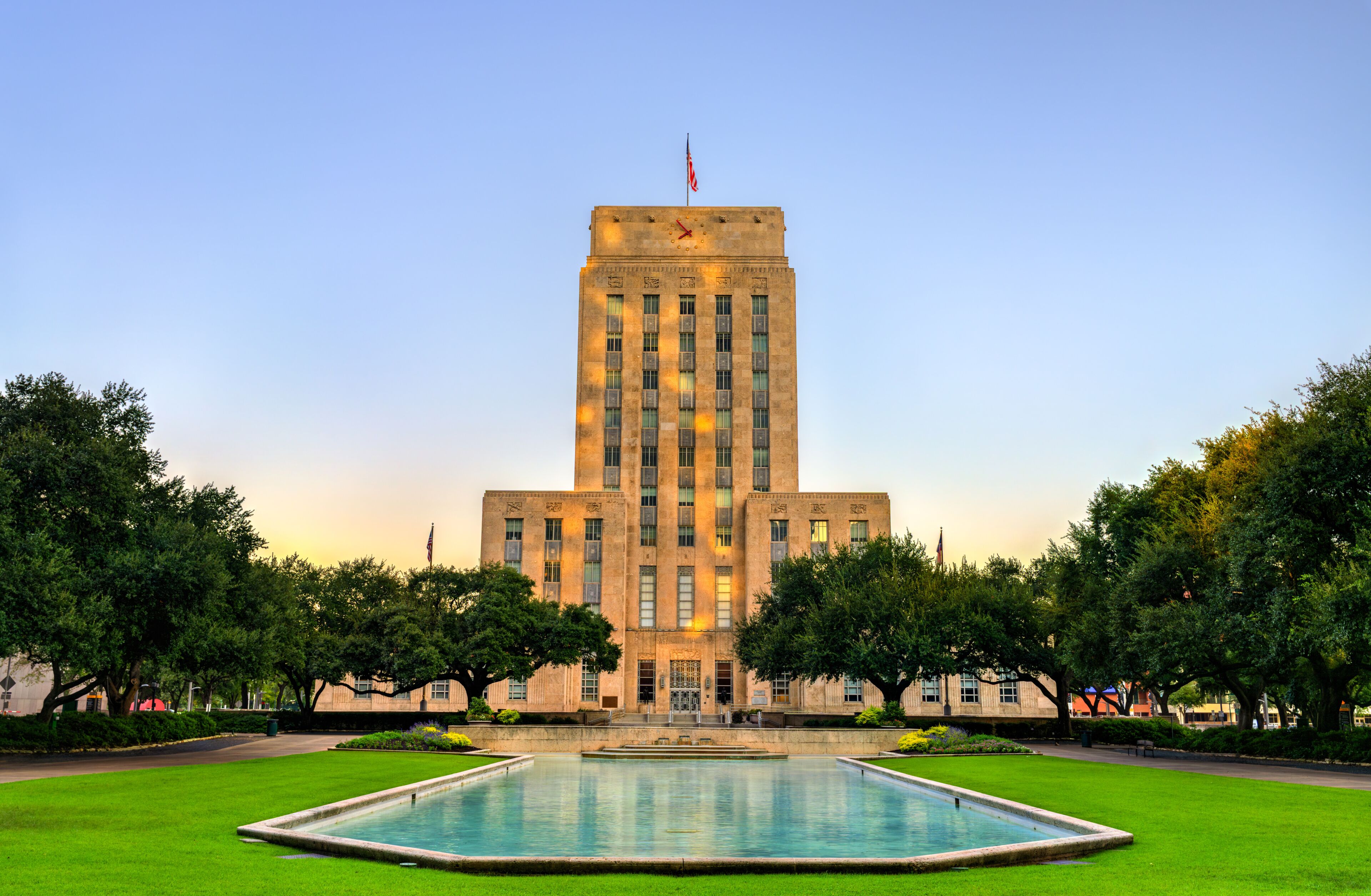 Houston City Hall viewed from Hermann Square, illuminated by warm sunrise light. The reflecting pool in the foreground mirrors the clear sky