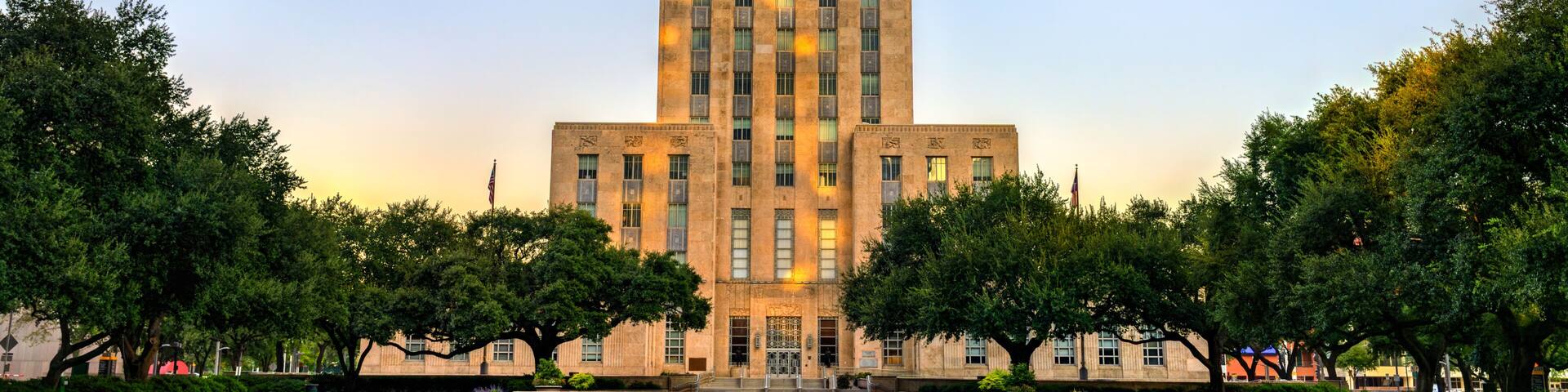 Houston City Hall viewed from Hermann Square, illuminated by warm sunrise light. The reflecting pool in the foreground mirrors the clear sky