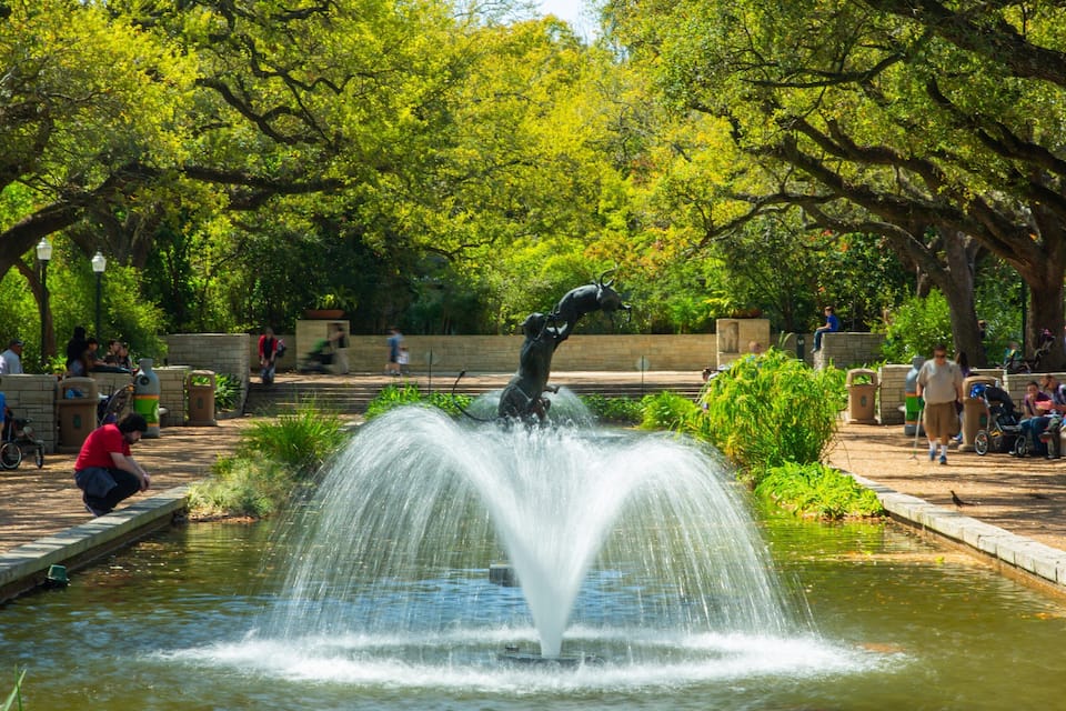 A park with a water fountain, trees, and people.