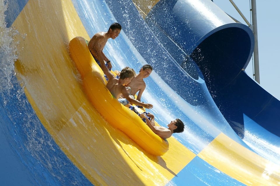 A water slide with yellow and blue sections, a large blue water jet, and people on a yellow raft.