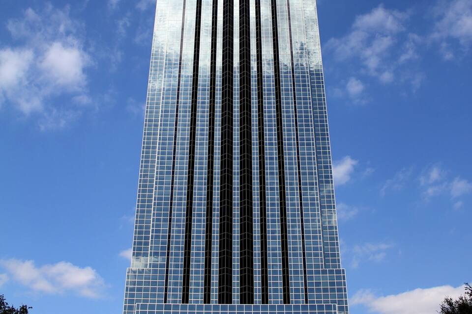 A tall, modern skyscraper with reflective glass windows and vertical lines, set against a clear blue sky with scattered clouds.