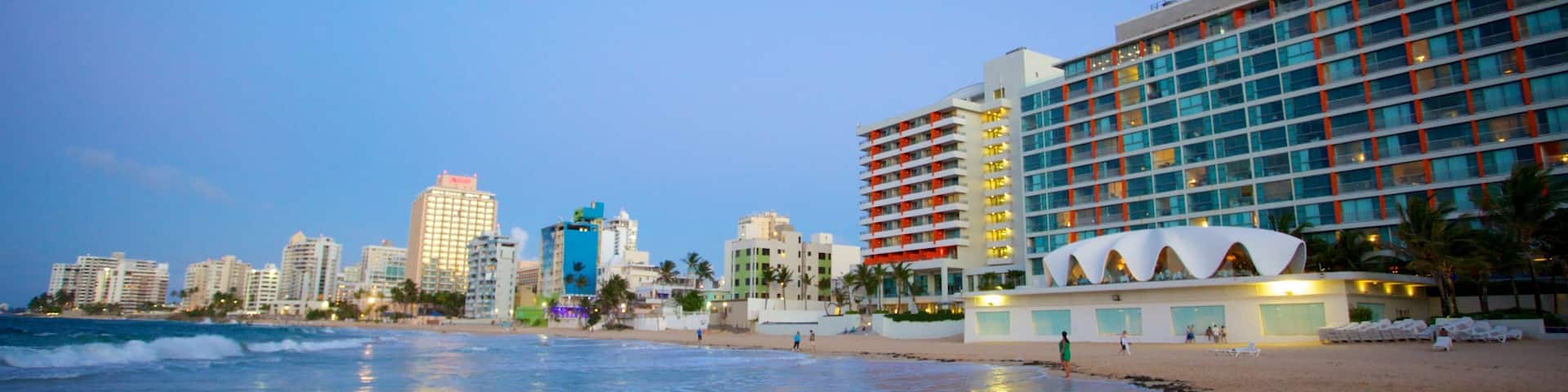A beach with ocean waves, a city skyline, and a hotel with a distinctive white and orange facade.