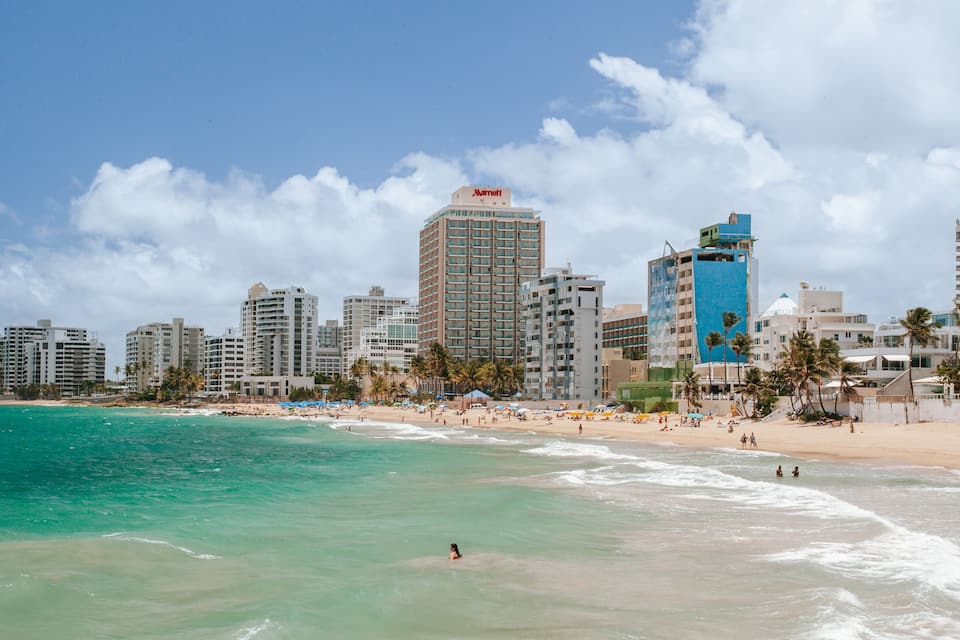A beach with clear turquoise water, a sandy shore, and a city skyline with tall buildings including a Marriott hotel.