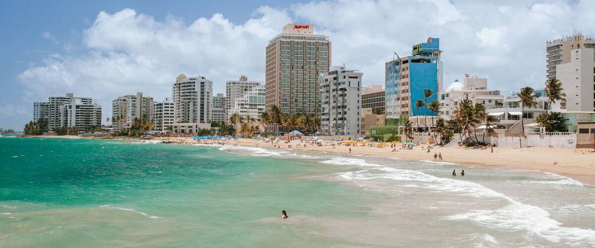 A beach with clear turquoise water, a sandy shore, and a city skyline with tall buildings including a Marriott hotel.