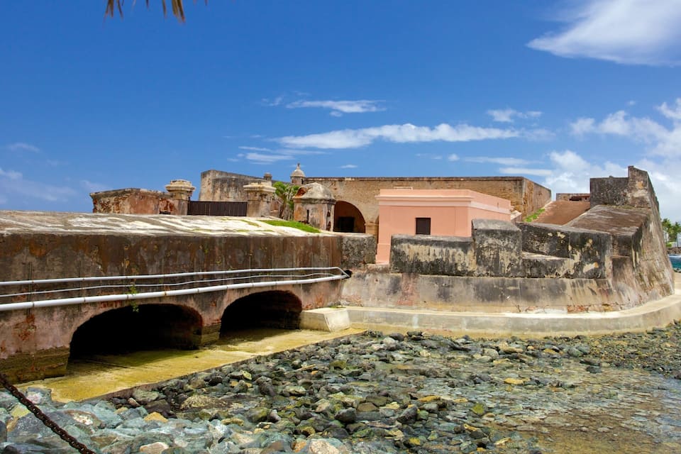 Una fortaleza costera con un puente, playa rocosa y cielo azul despejado.