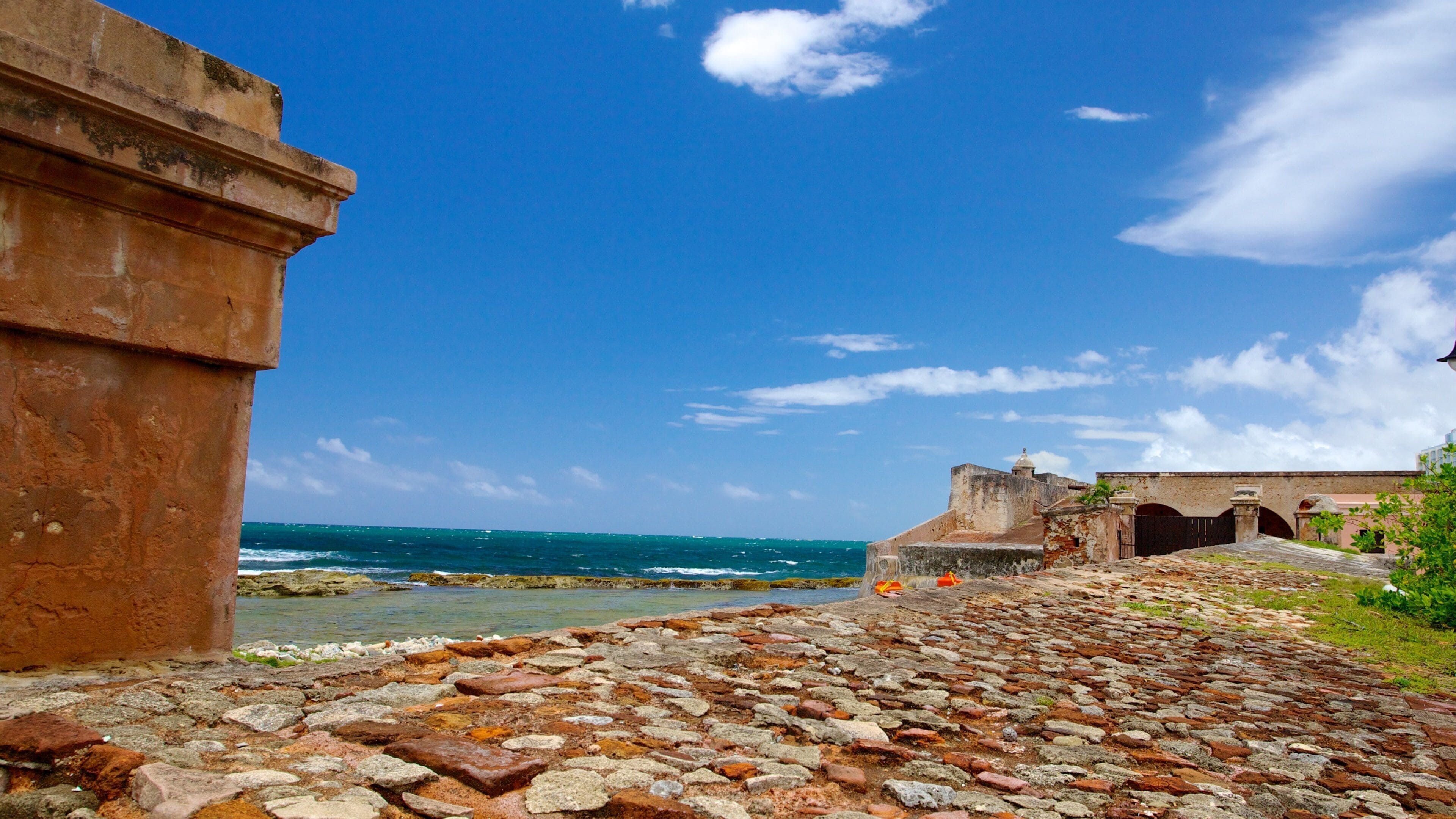 Una fortificación costera con un sendero adoquinado que lleva al mar, bajo un cielo azul despejado.