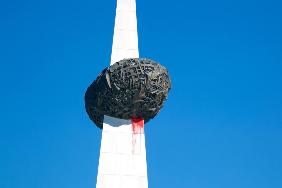 A large, intricate sculpture mounted on a tall white pole against a clear blue sky.