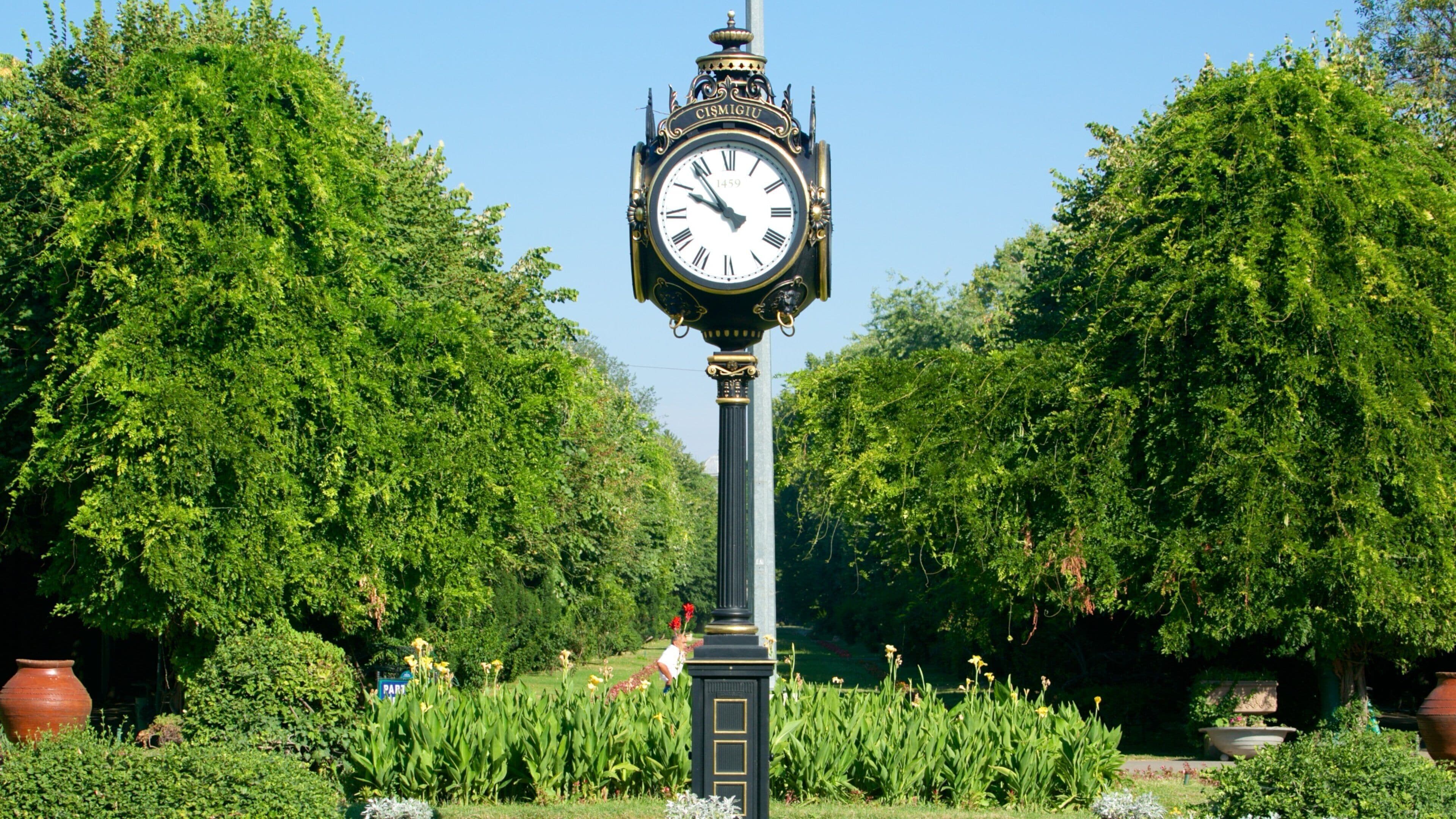 Una torre de reloj con esfera de números romanos, rodeada de vegetación exuberante y árboles.