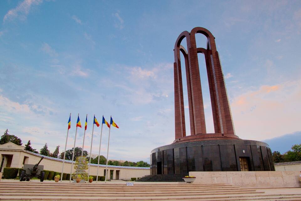 A monument with three tall arches, surrounded by steps and flags.