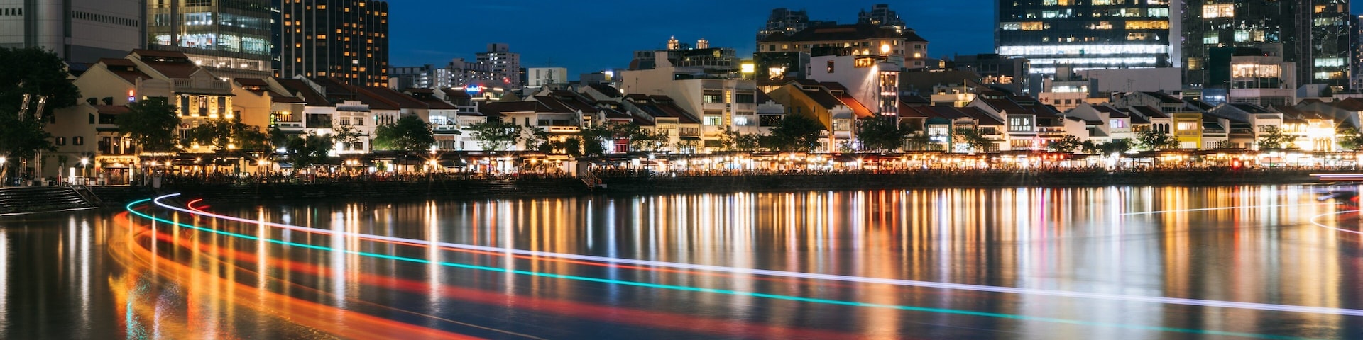 Paisagem urbana noturna com edifícios iluminados e um rio refletindo rastros de luz colorida.
