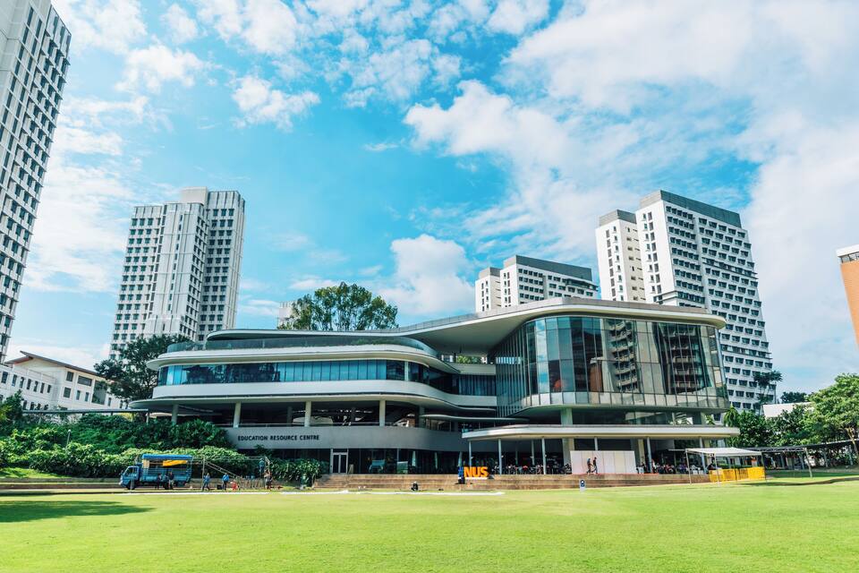 A modern educational building with a curved roof, surrounded by tall residential buildings and a green park.