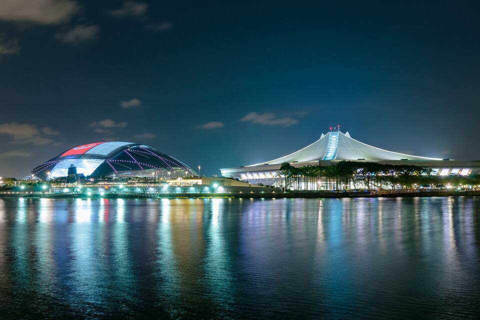 A stadium with a distinctive red and blue roof and a white building with a sloped roof near a body of water.