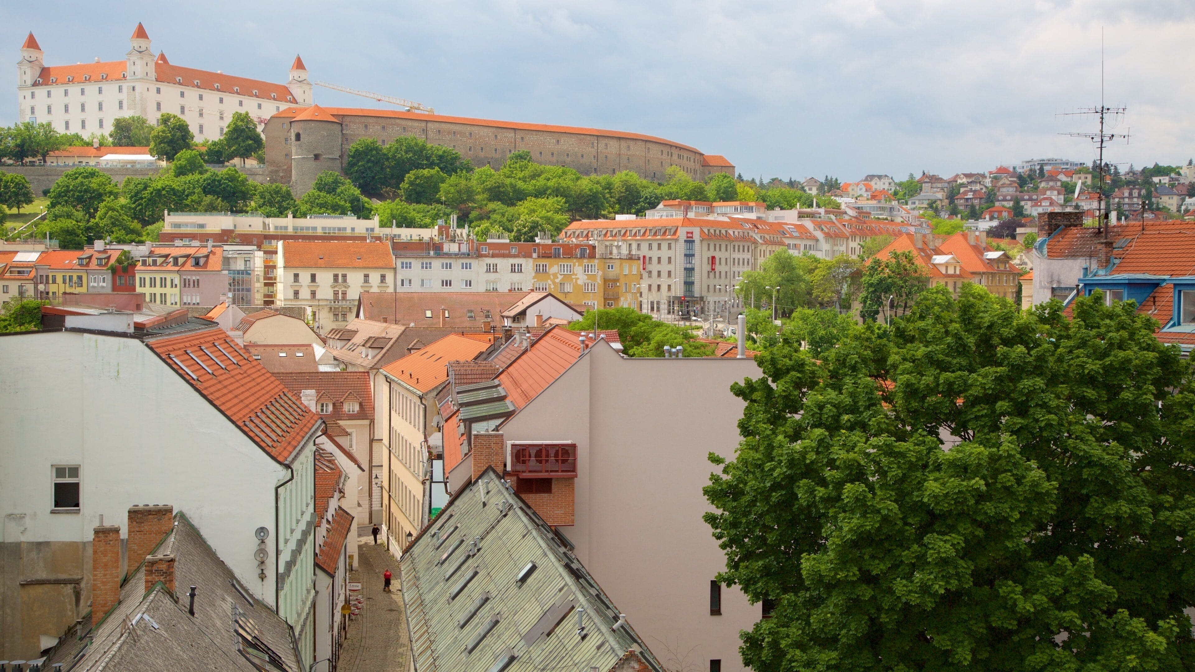 A historic castle with red-tiled roofs, surrounded by colorful buildings and lush green trees.