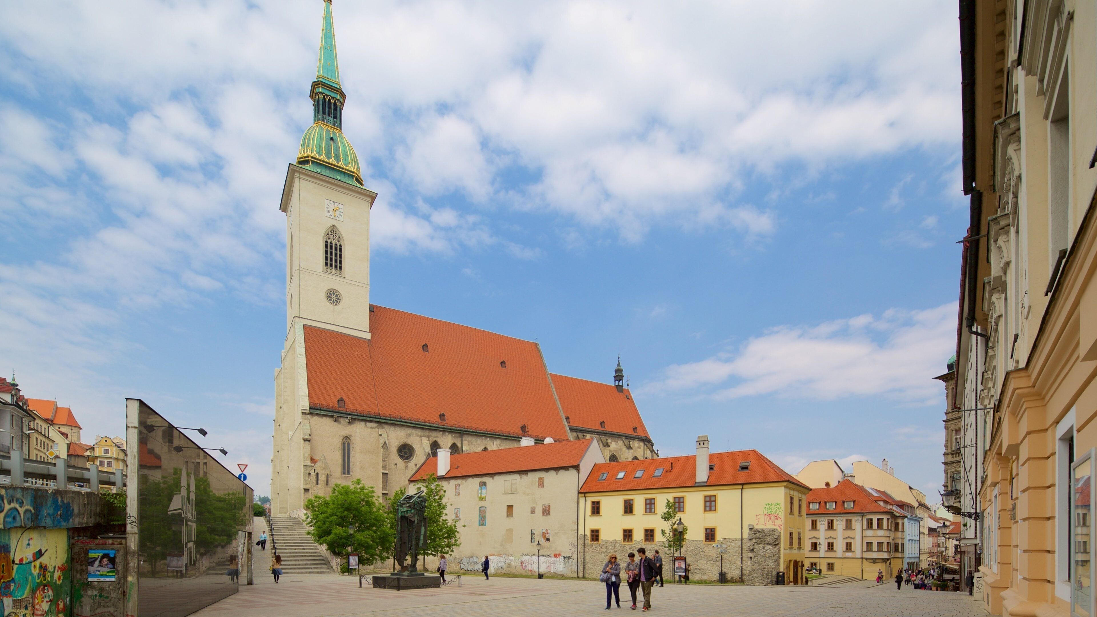 A historic church with a tall tower and a red roof, surrounded by old buildings and a cobblestone square.