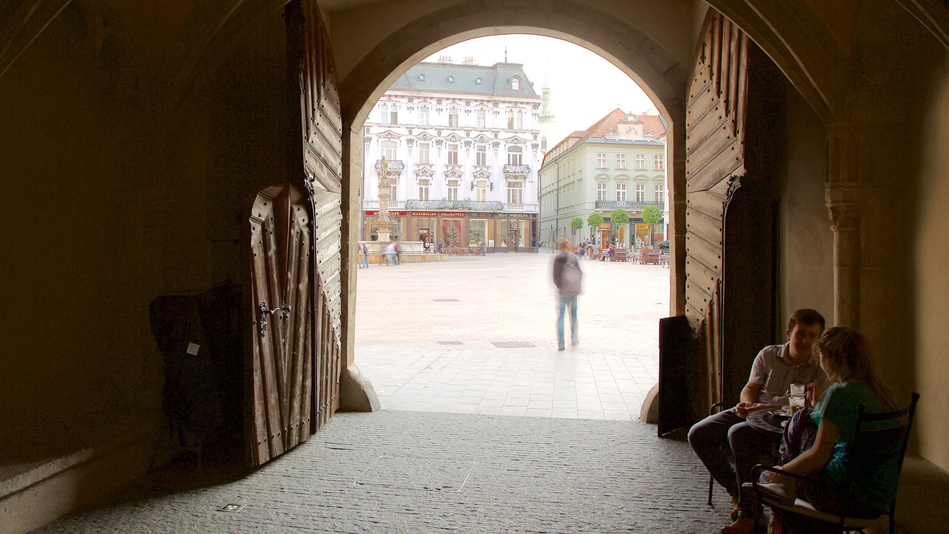 An open archway leading to a square with buildings and a statue.