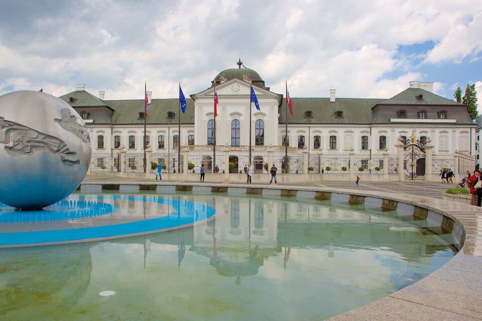 A large spherical sculpture in a reflective pool, a historic building with flags, and people walking around.