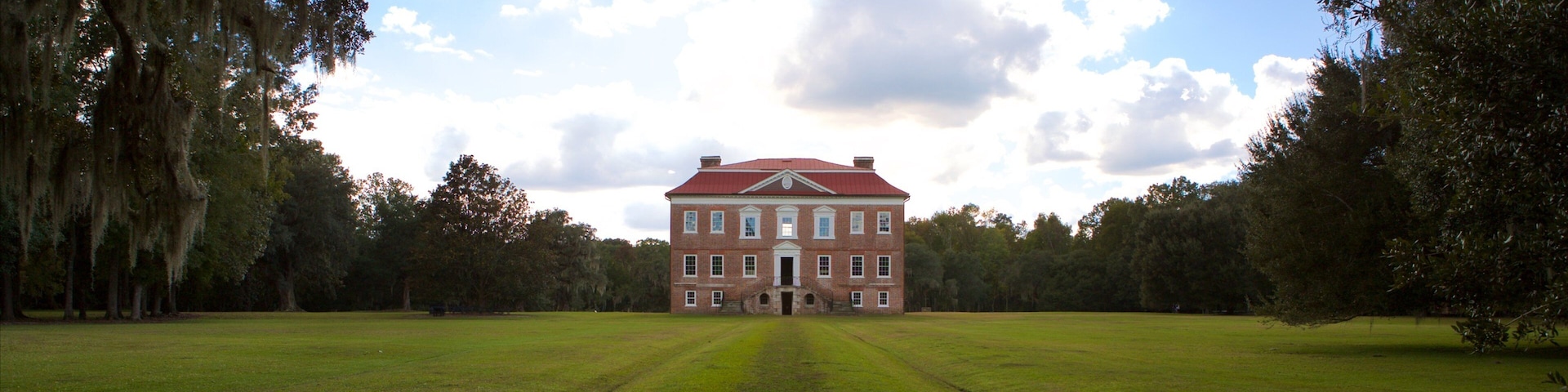 Um grande edifício de tijolos com telhado vermelho, cercado por um campo gramado e árvores.