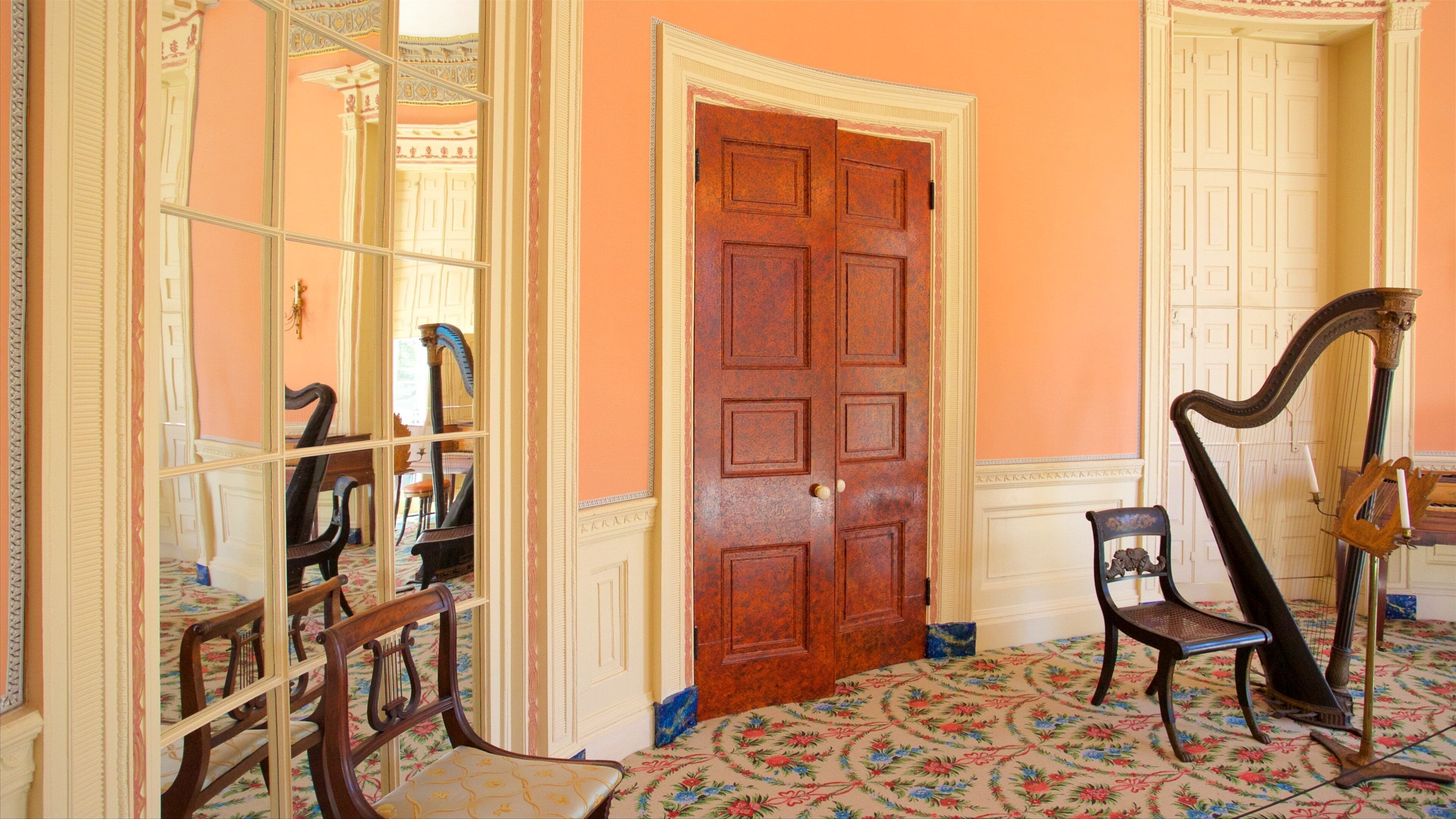 A room with a large wooden door, a harp, and a mirror reflecting a room with a piano.