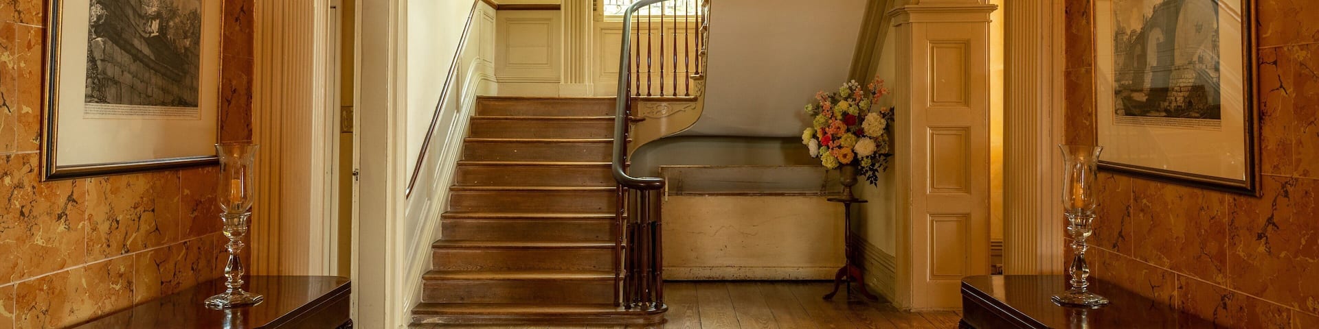 A grand staircase with a wooden finish, flanked by framed pictures and a vase of flowers.