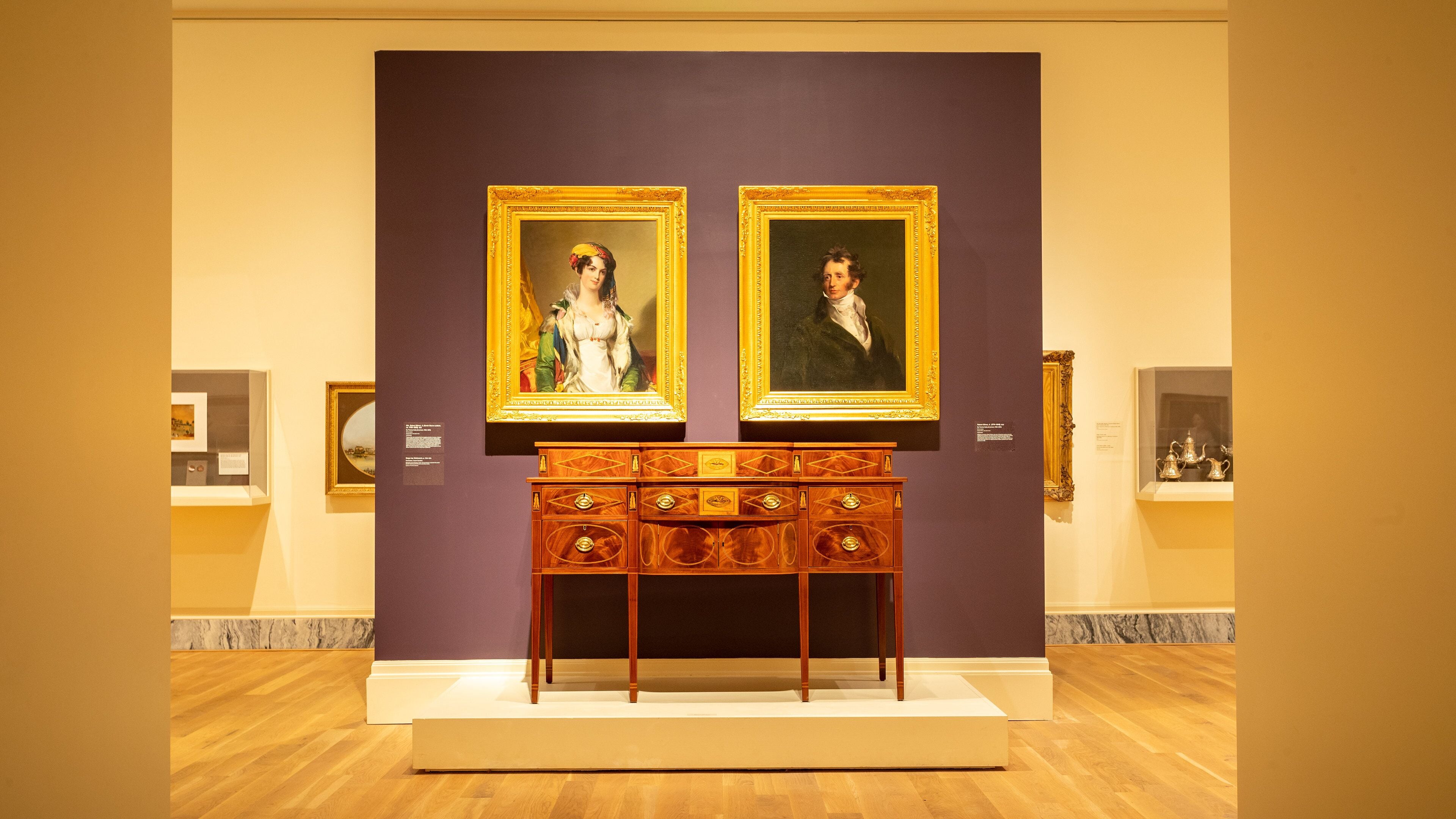 A museum exhibit with two framed paintings on a dark purple wall, a wooden cabinet with drawers, and marble flooring.