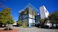 A modern glass building with a unique geometric design, surrounded by trees and a paved plaza.