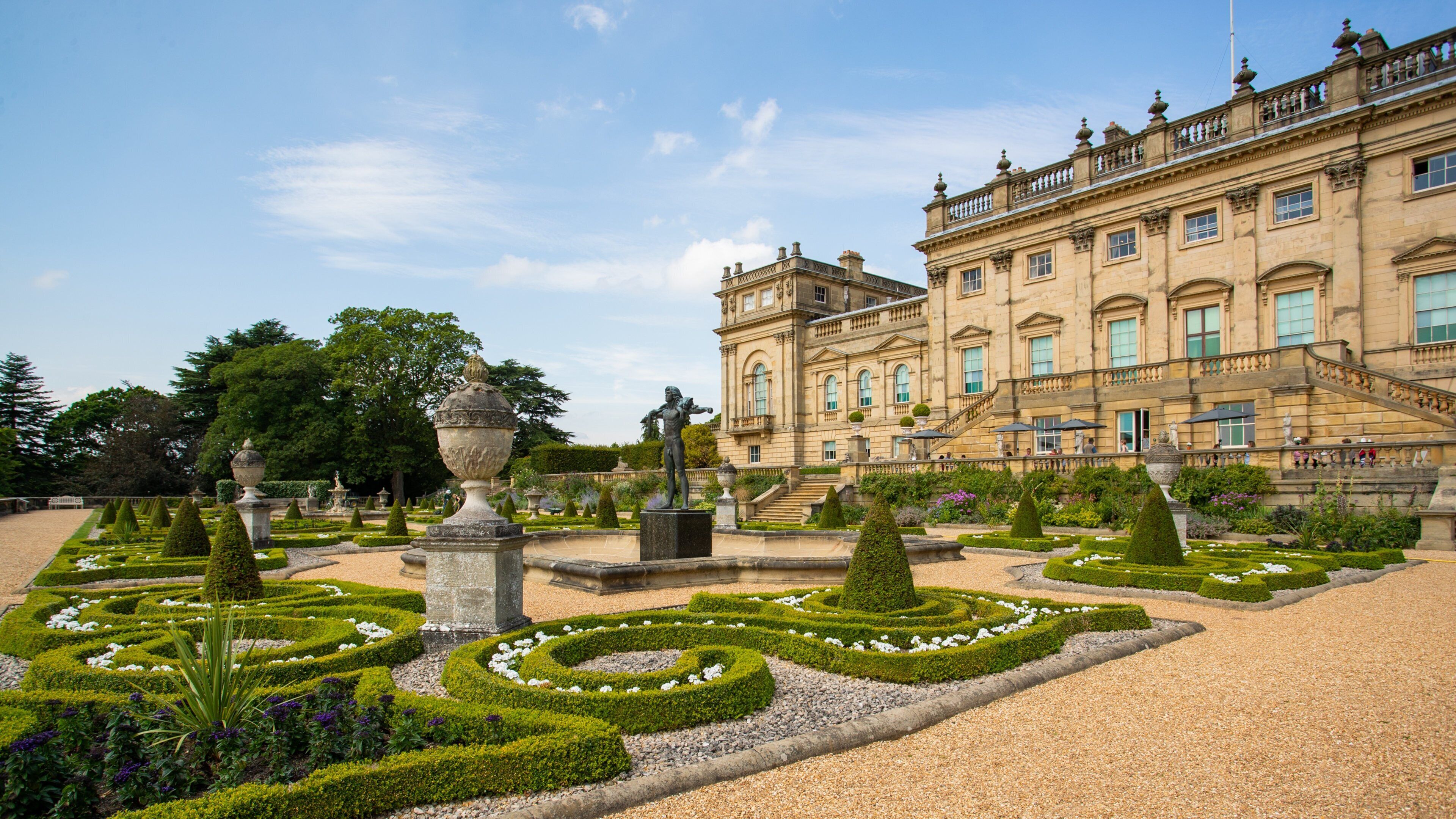 A grand historic building with formal gardens, including trimmed hedges and statues.